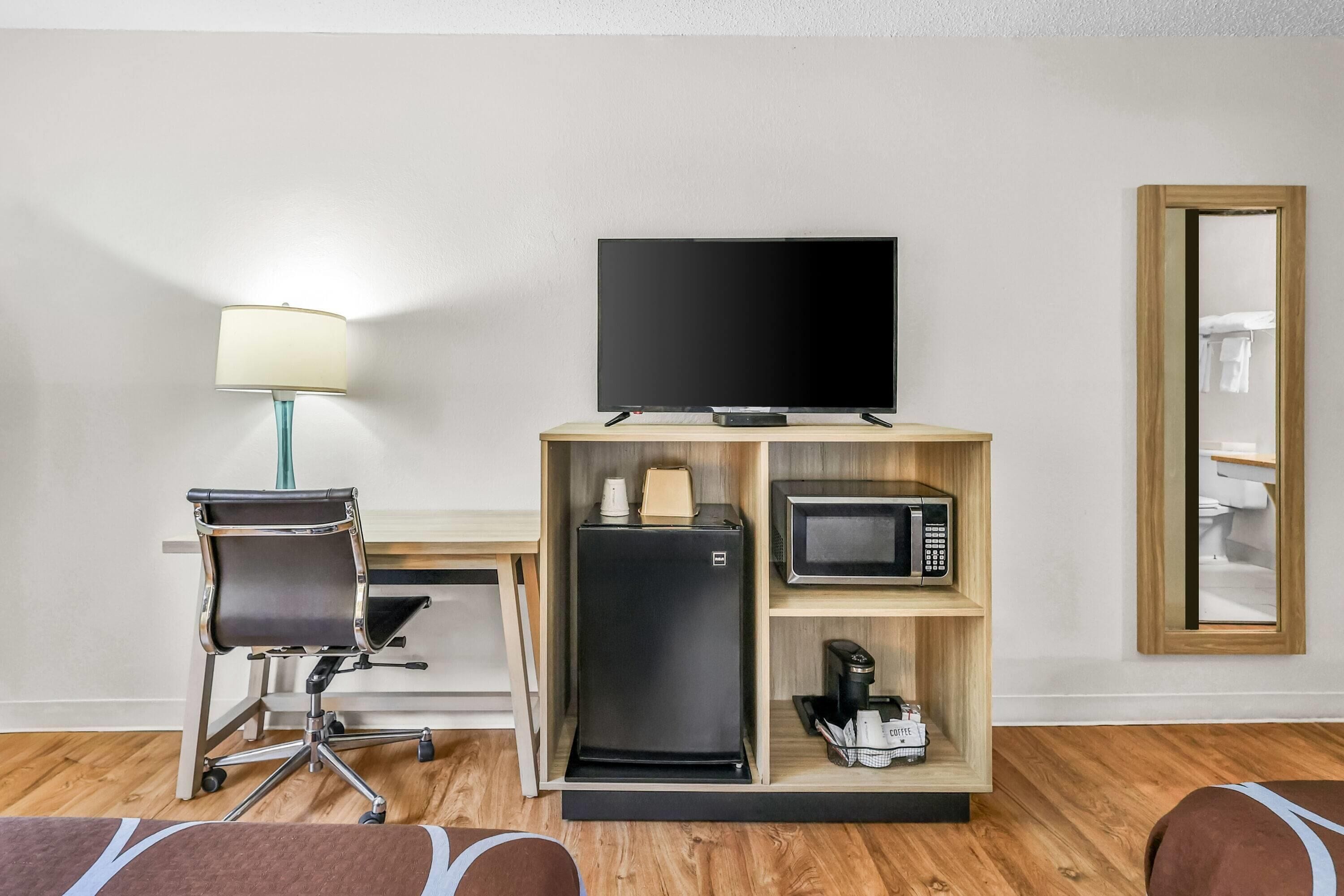 A hotel room with a desk, chair, television, microwave, refrigerator, and a mirror.