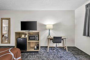 Habitación de hotel con cama, escritorio con silla, televisor, refrigerador, microondas, lámpara y espejo.