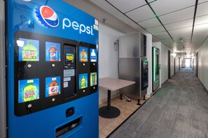 A vending machine with various beverage options, a table, and a corridor with doors and a refrigerator. - Red Roof Inn Greensburg (Greensburg)