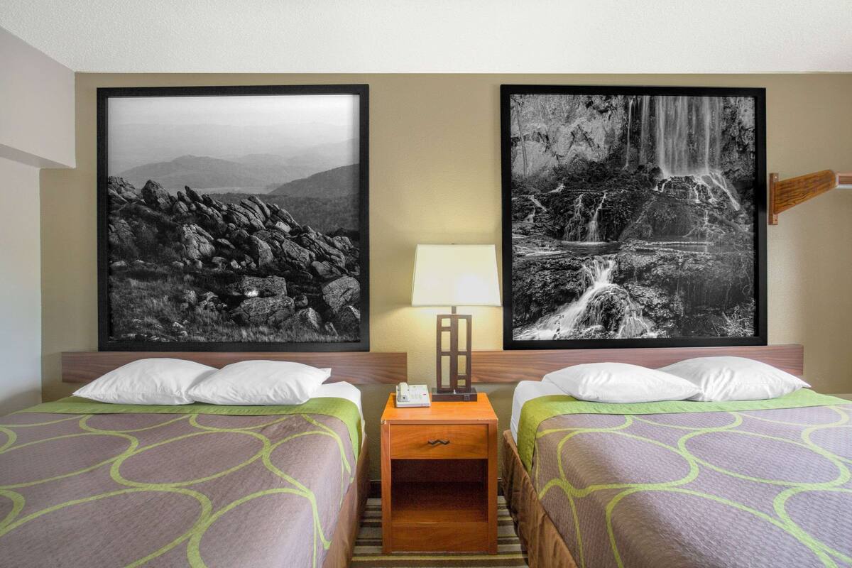 A hotel room with two beds, a nightstand, a lamp, and two framed black and white photographs on the wall.