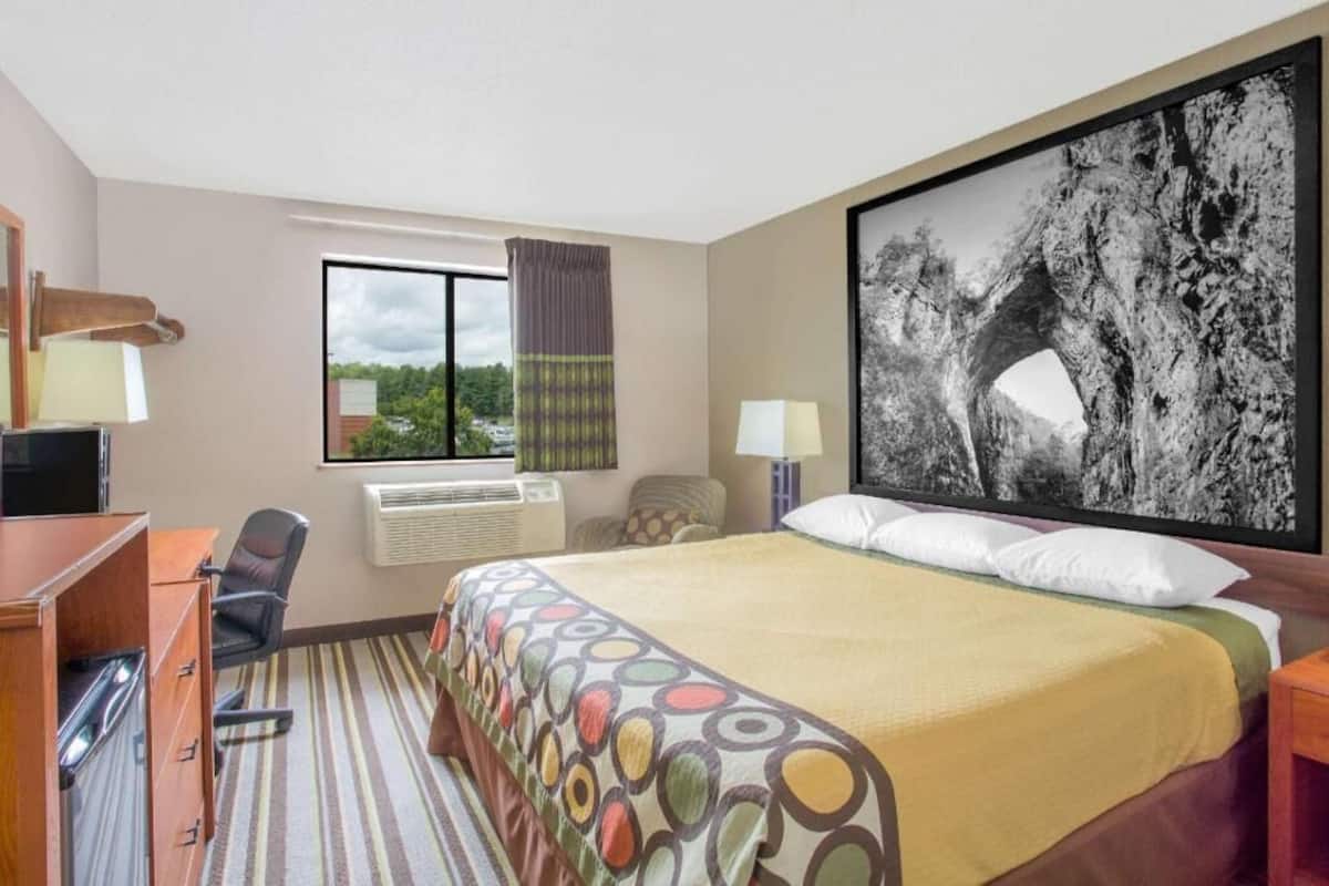 Ein Hotelzimmer mit Bett, Schreibtisch mit Computer, Stuhl, Klimaanlage, Fenster mit Vorhängen und einem großen Bild in einem Rahmen an der Wand.