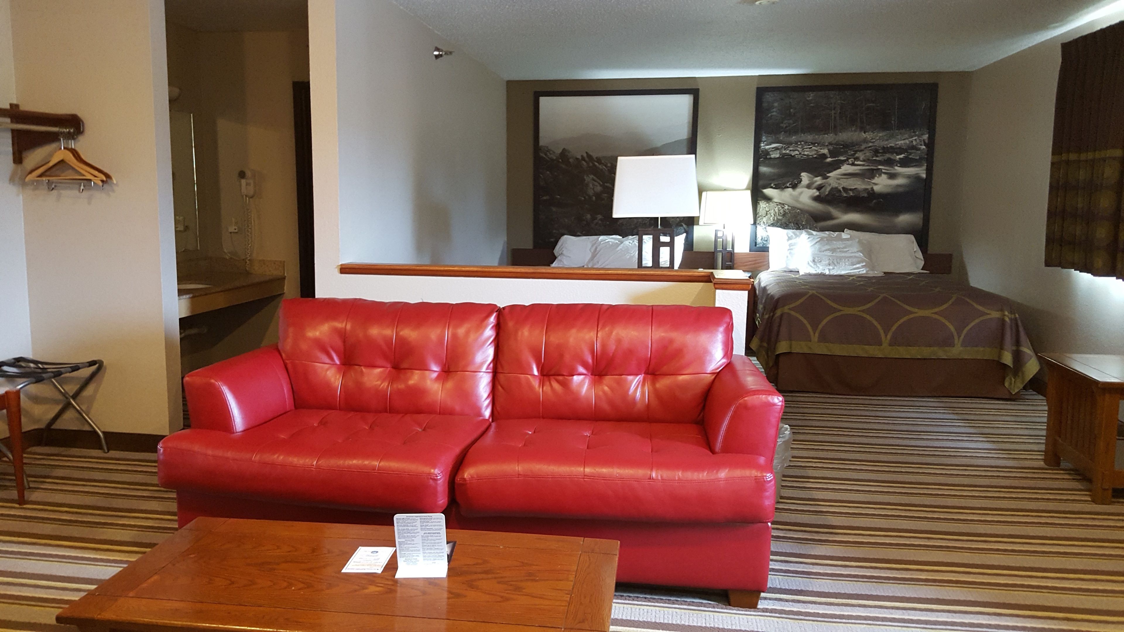 A hotel room with a red leather sofa, a wooden coffee table, a bed with a patterned bedspread, a nightstand, and a large window with a view of a mountain landscape.