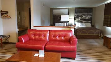 A hotel room with a red leather sofa, a wooden coffee table, a bed with a patterned bedspread, a nightstand, and a large window with a view of a mountain landscape.