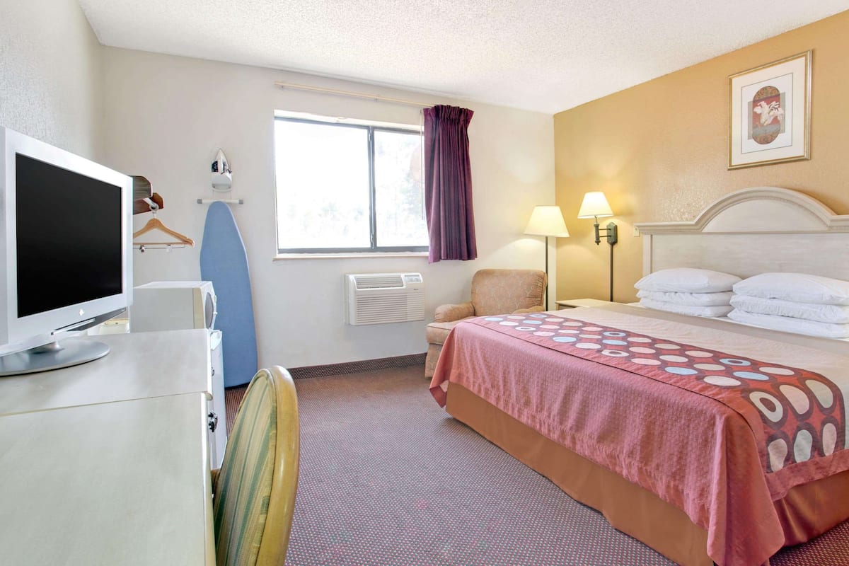 Ett hotellrum med en säng, ett skrivbord med en dator, en strykbräda, en stol, ett fönster med gardiner och en vägglampa.