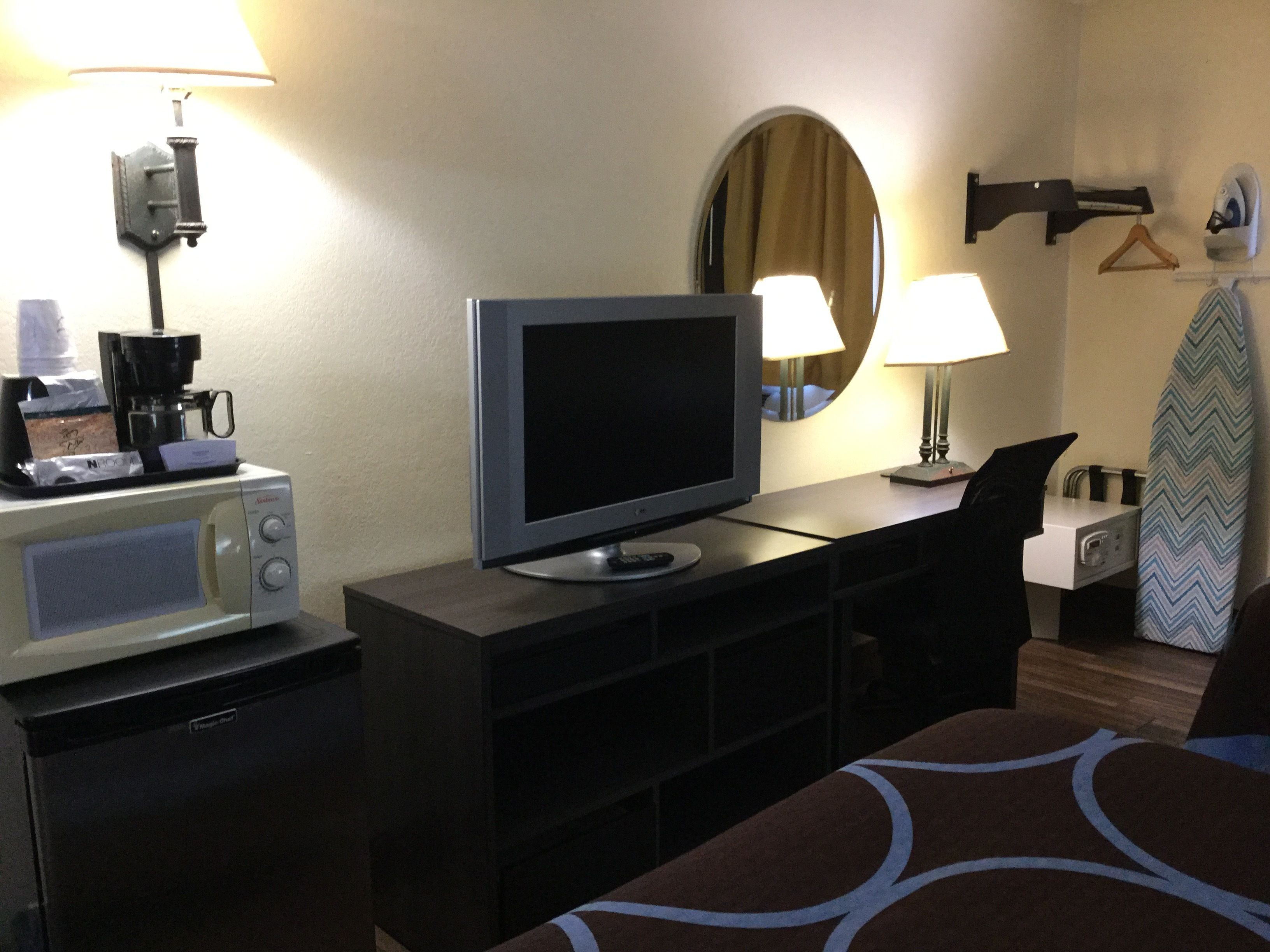 A hotel room with a television, a coffee maker, a desk, a chair, a bed, a mirror, and a coat rack with a hanger.