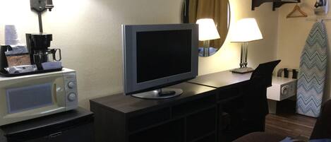 A hotel room with a television, a coffee maker, a desk, a chair, a bed, a mirror, and a coat rack with a hanger.
