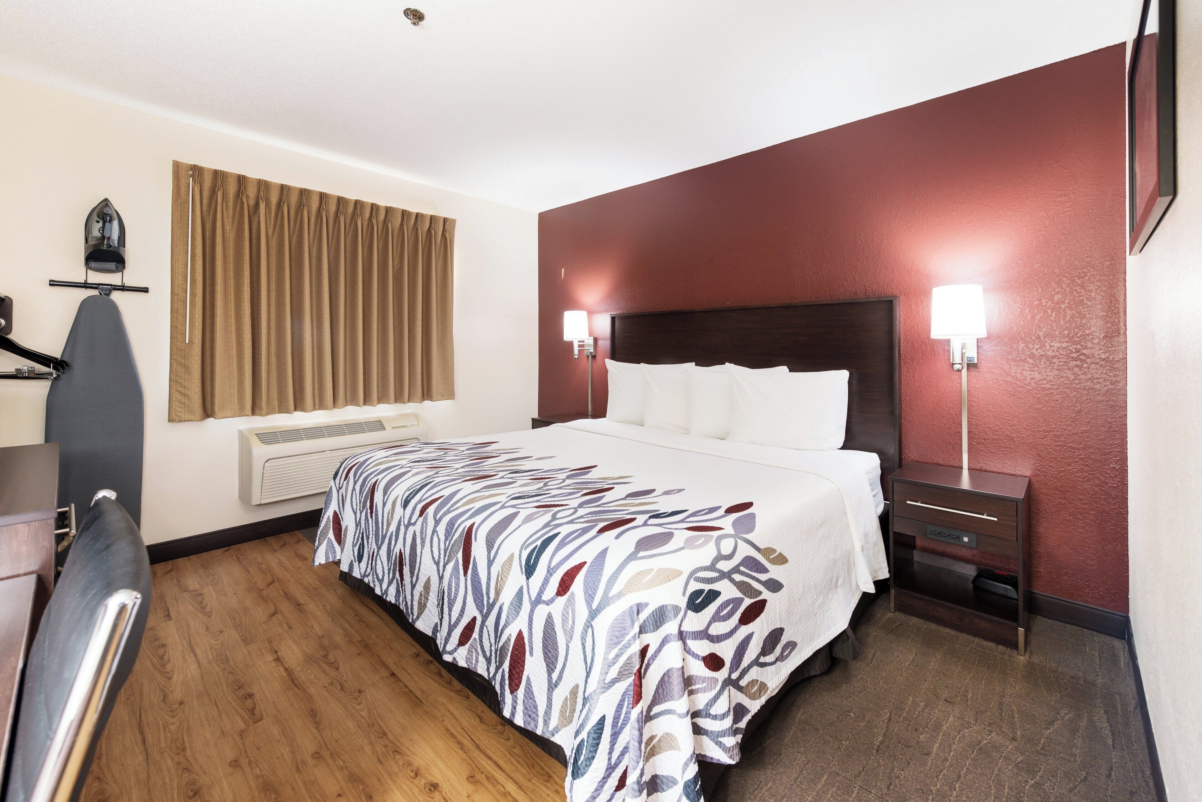 A hotel room with a bed, a desk with an ironing board, a nightstand, and a wall-mounted lamp.