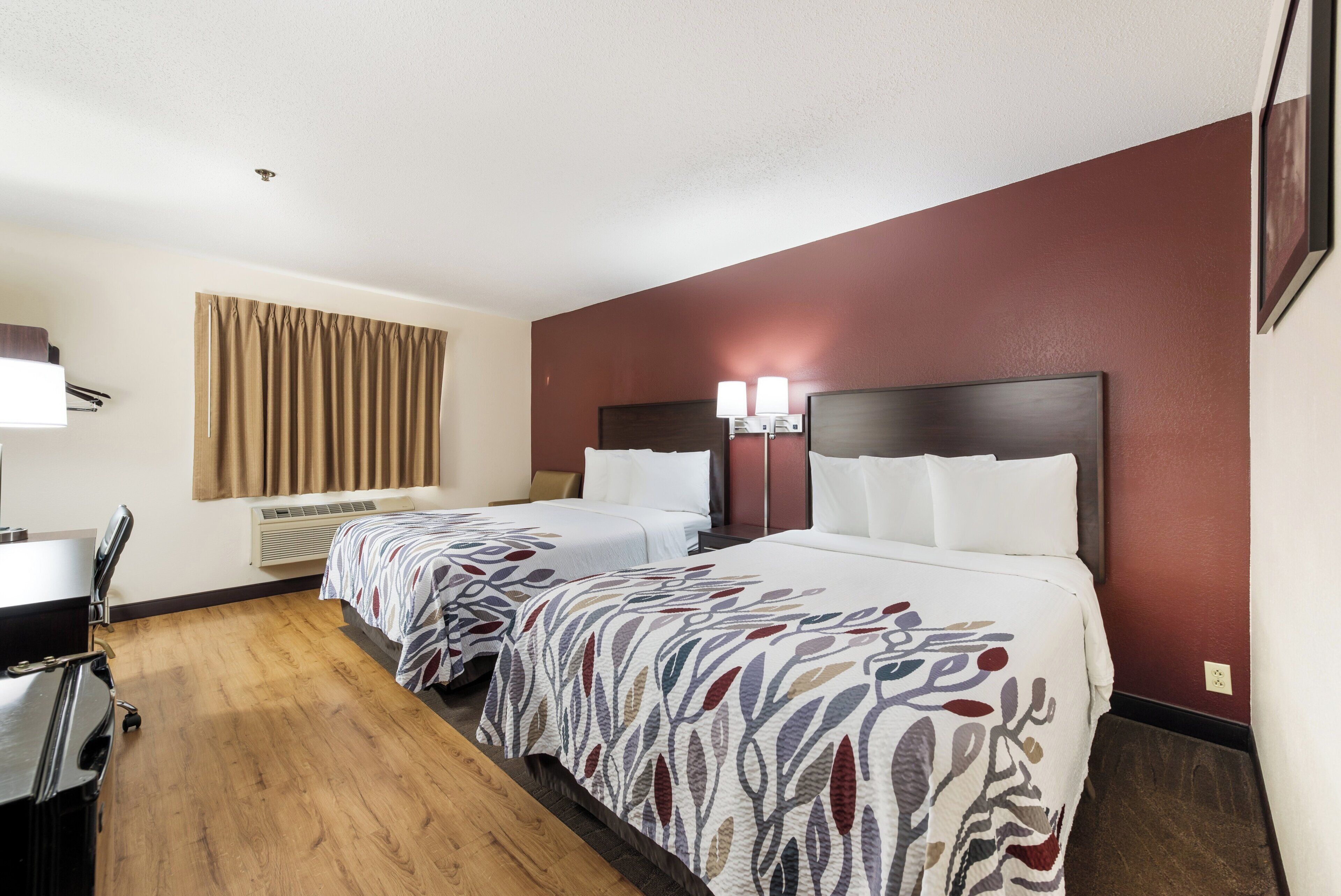A hotel room with two beds, a desk, a chair, a window with curtains, and a wall-mounted lamp.