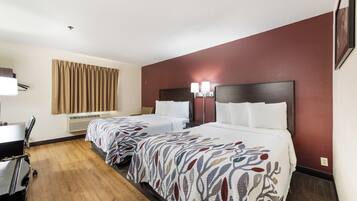 A hotel room with two beds, a desk, a chair, a window with curtains, and a wall-mounted lamp.
