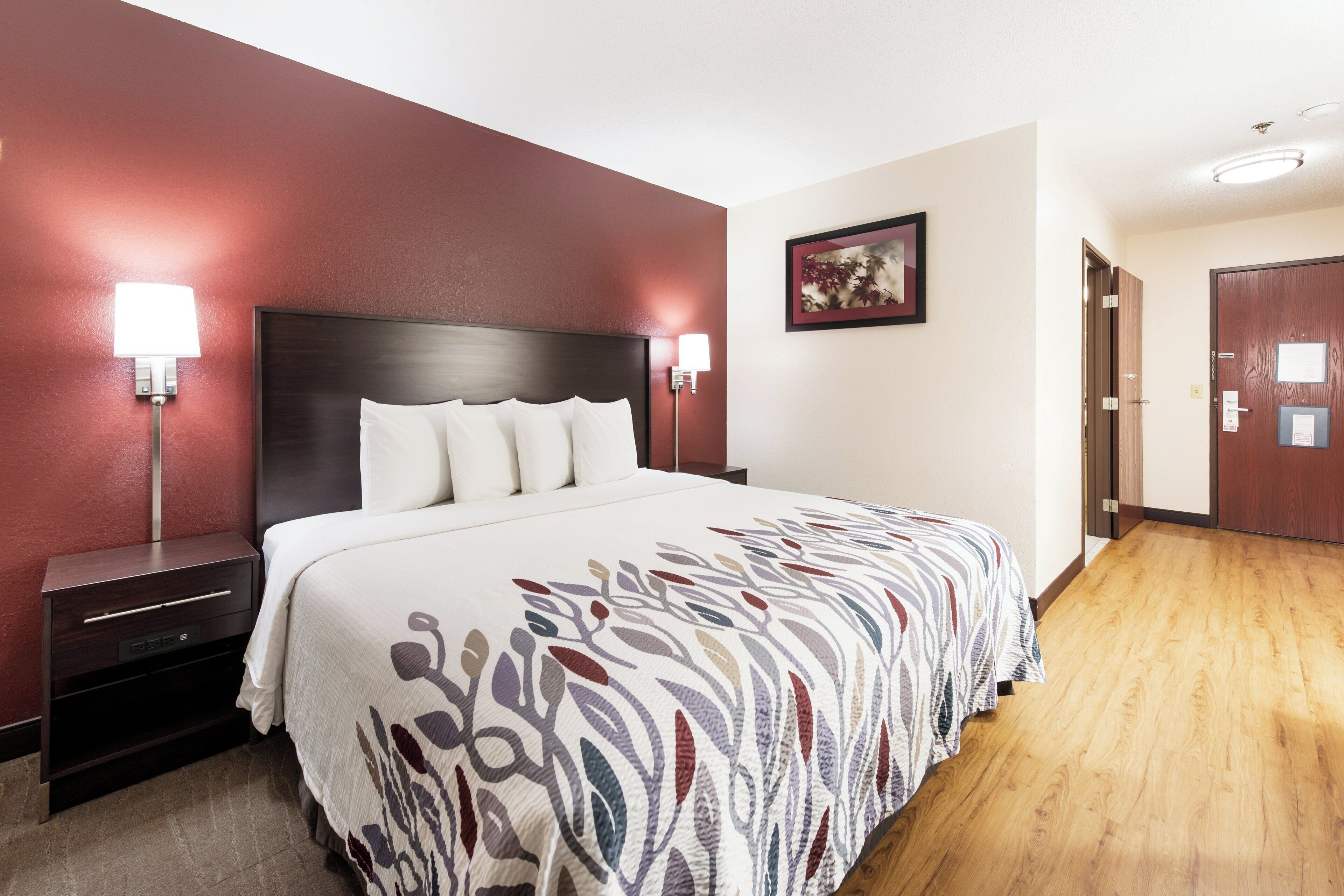 A hotel room with a large bed, two bedside tables, a nightstand, a wall-mounted lamp, a framed picture, and a door.