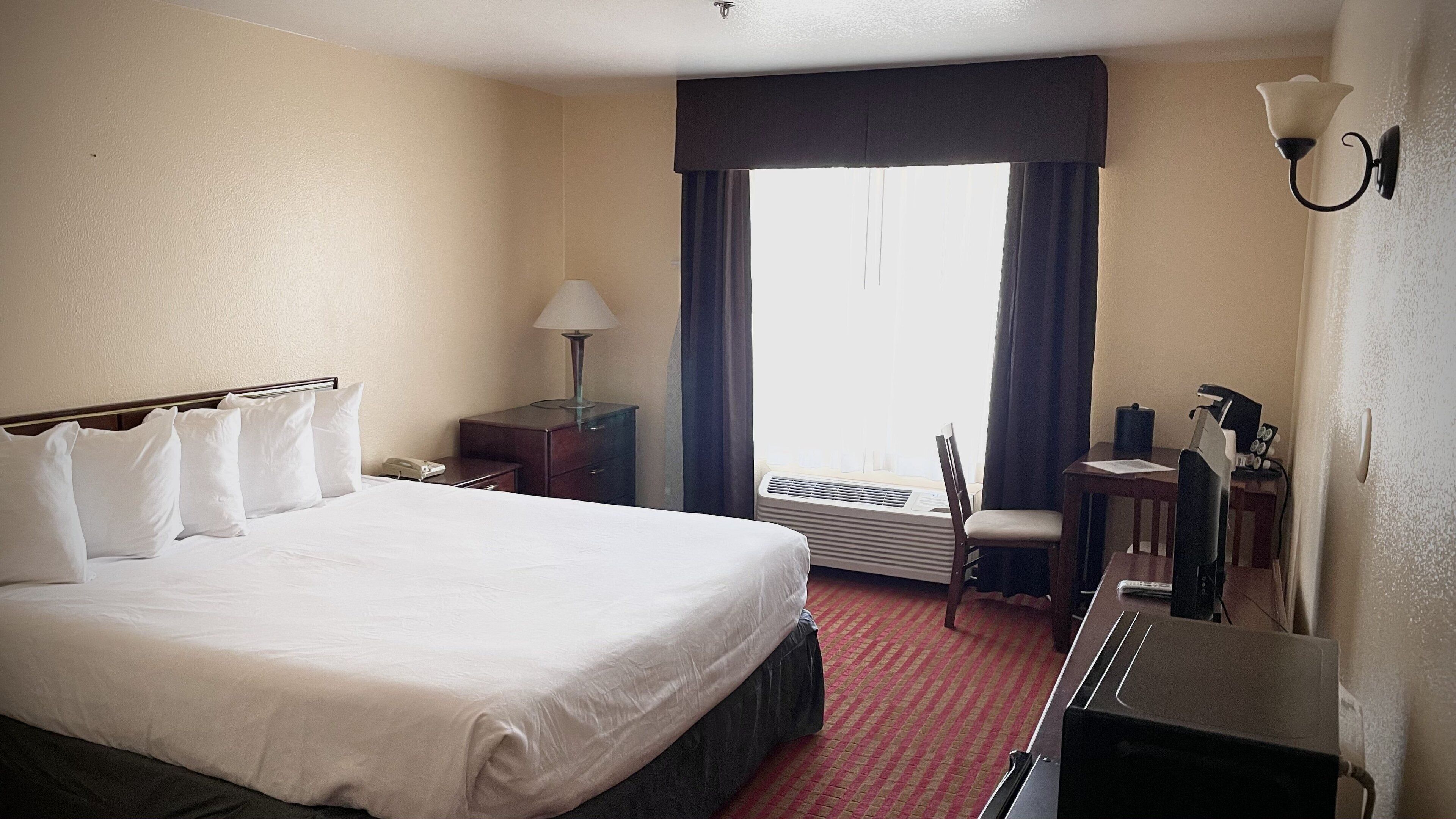 A hotel room with a large bed, a desk with a chair, a lamp, and an air conditioning unit.