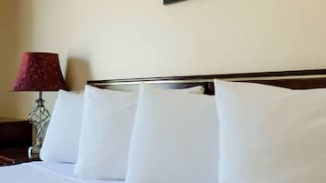 Una cama bien hecha con sábanas y almohadas blancas, un cuadro en la pared y una lámpara con pantalla estampada sobre una mesita de noche.