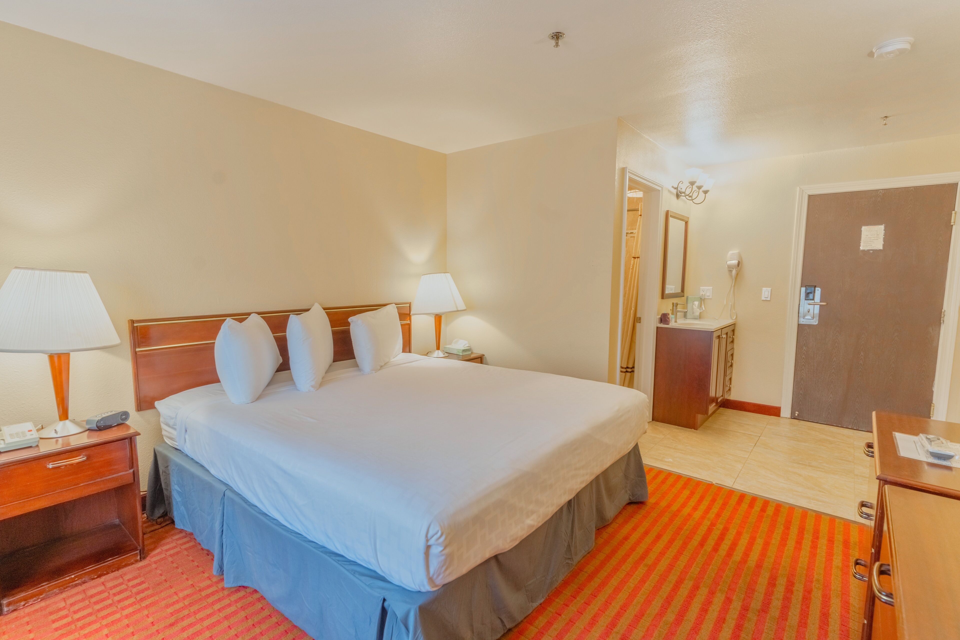 Una habitación de hotel con una cama grande, lámparas de noche, una mesita de noche de madera, un escritorio y una puerta.