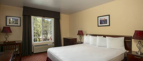 A hotel room with a large bed, two bedside tables with lamps, a desk with a chair, a window with curtains, and two framed pictures on the walls.