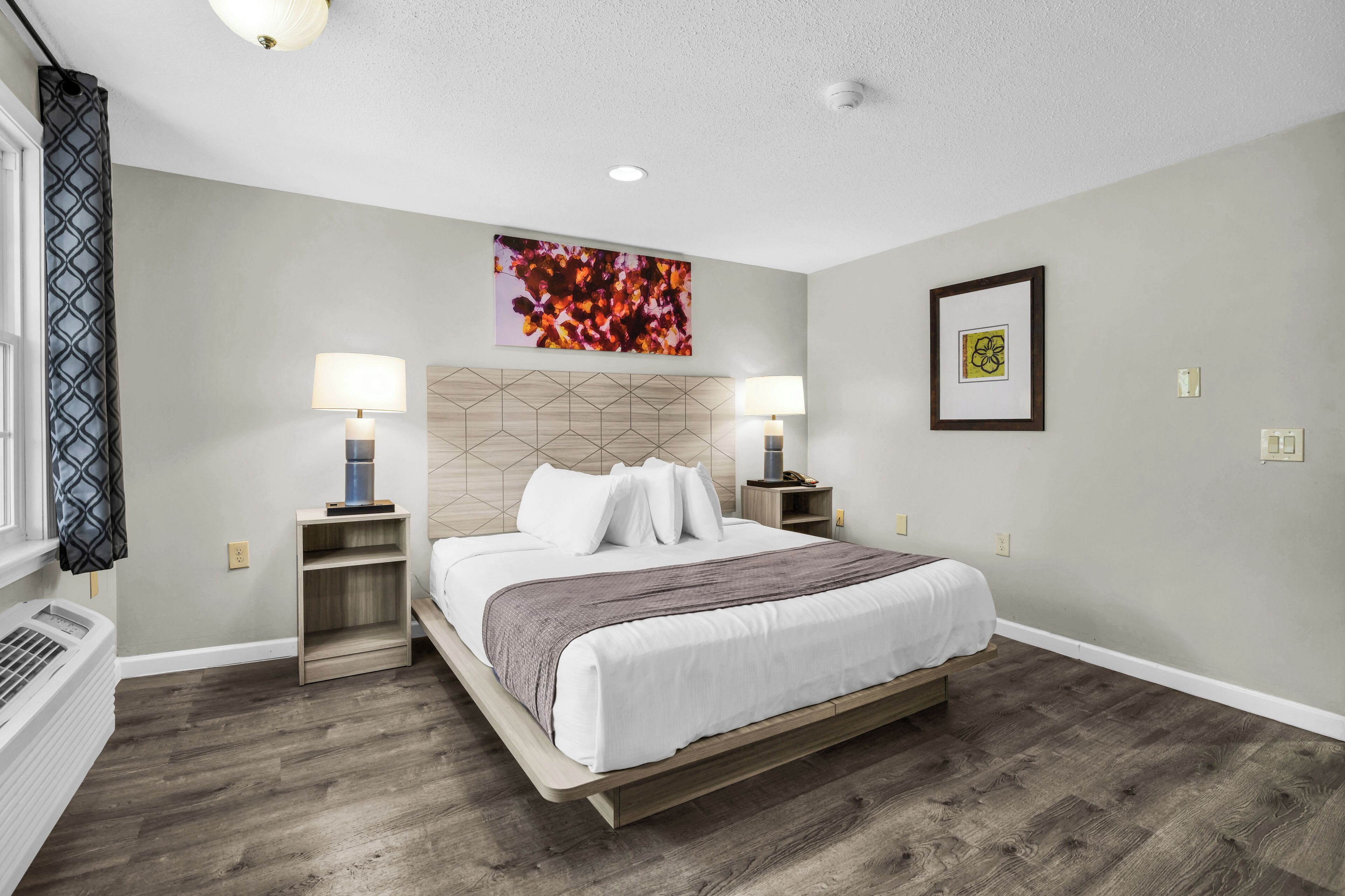 A bedroom with a bed, two bedside tables, a heater, a window with curtains, and a wall with a framed picture.