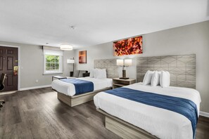 A hotel room with two beds, a desk, a chair, a window, and artwork on the walls.