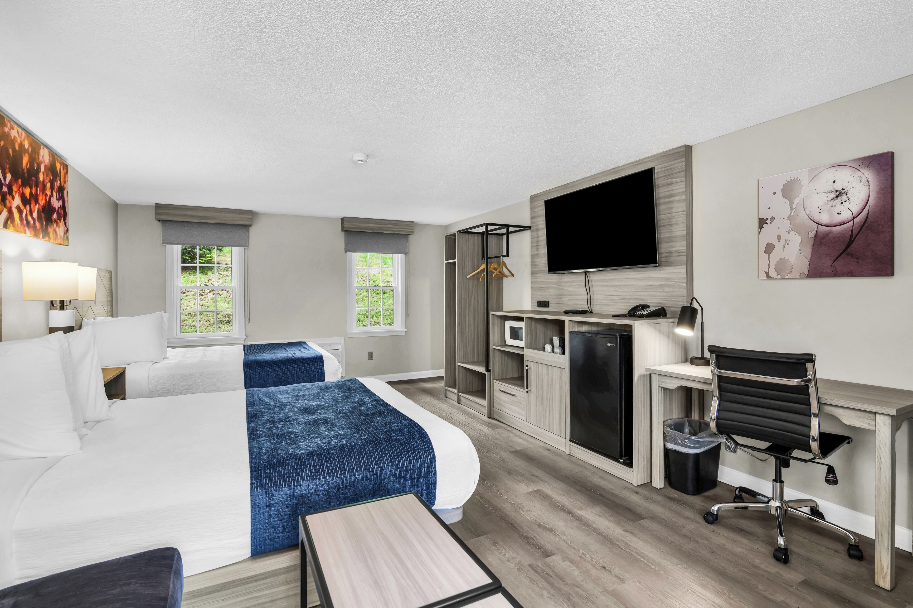 A hotel room with a bed, desk, chair, TV, and artwork.