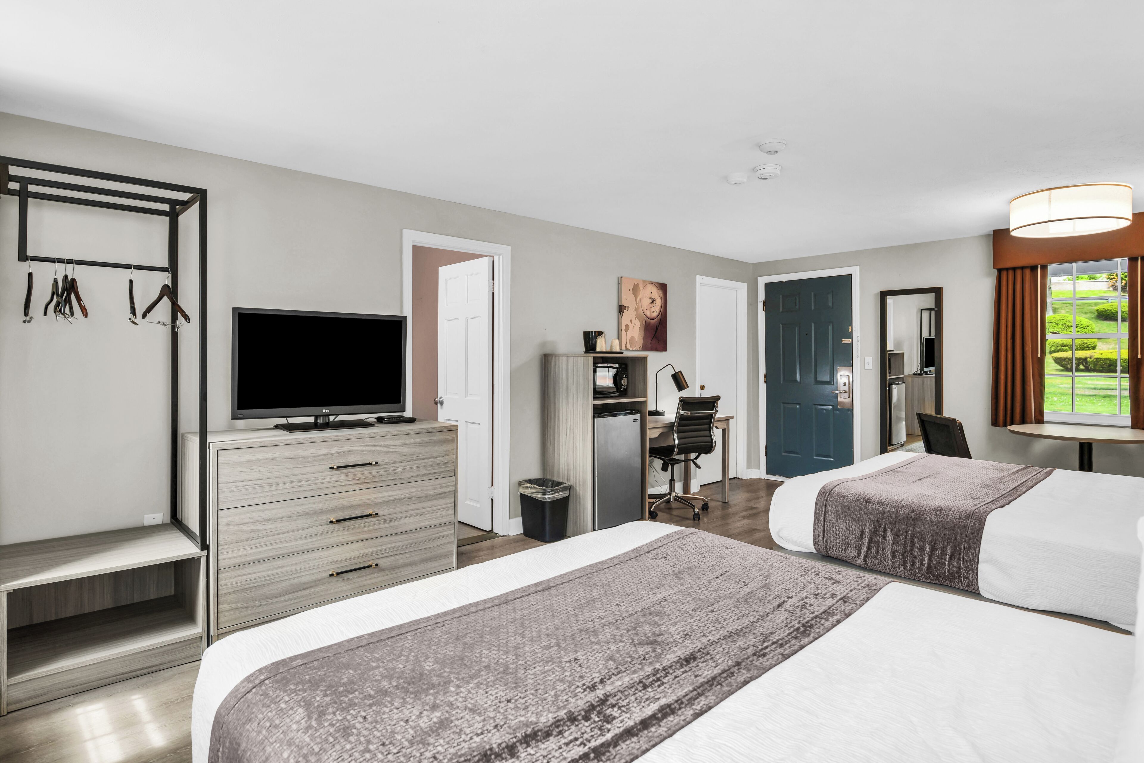 A hotel room with two beds, a TV, a desk with a computer, a mirror, and a view of a green landscape.