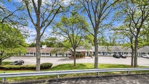 A motel with a parking lot, several trees, and a building with a sloped roof.
