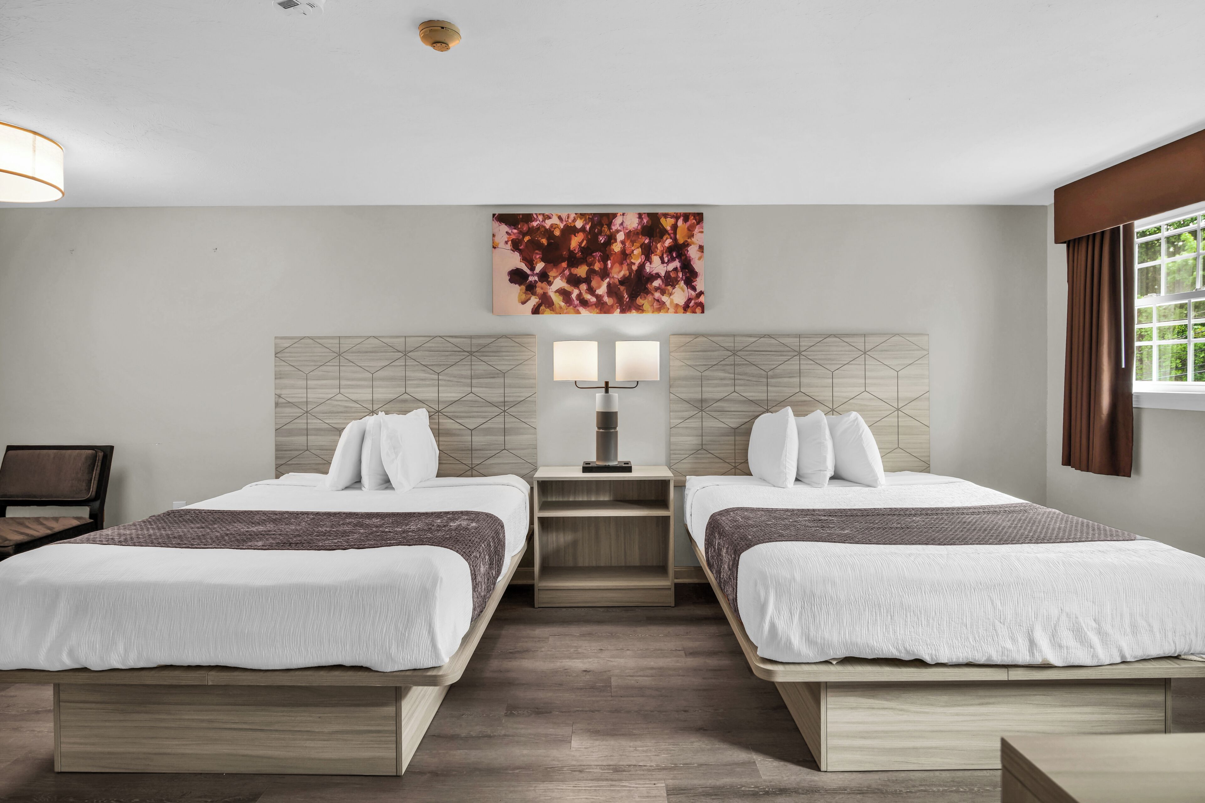 Une chambre d’hôtel avec deux lits, une table de chevet avec une lampe, une œuvre d’art géométrique accrochée au mur et une fenêtre avec des rideaux.