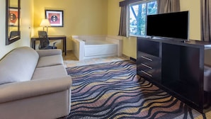 A hotel room with a sofa, a desk with a lamp, a flat-screen TV, a bathtub, and a patterned rug. - AmericInn by Wyndham Streator (Streator)