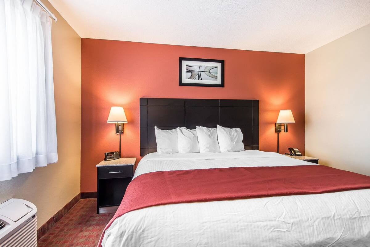 A hotel room with a large bed, two bedside tables with lamps, a framed picture on the wall, and a red throw blanket.