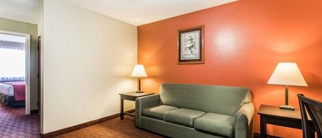 A hotel room with a patterned sofa, two bedside tables with lamps, a framed picture on the wall, and a carpeted floor.