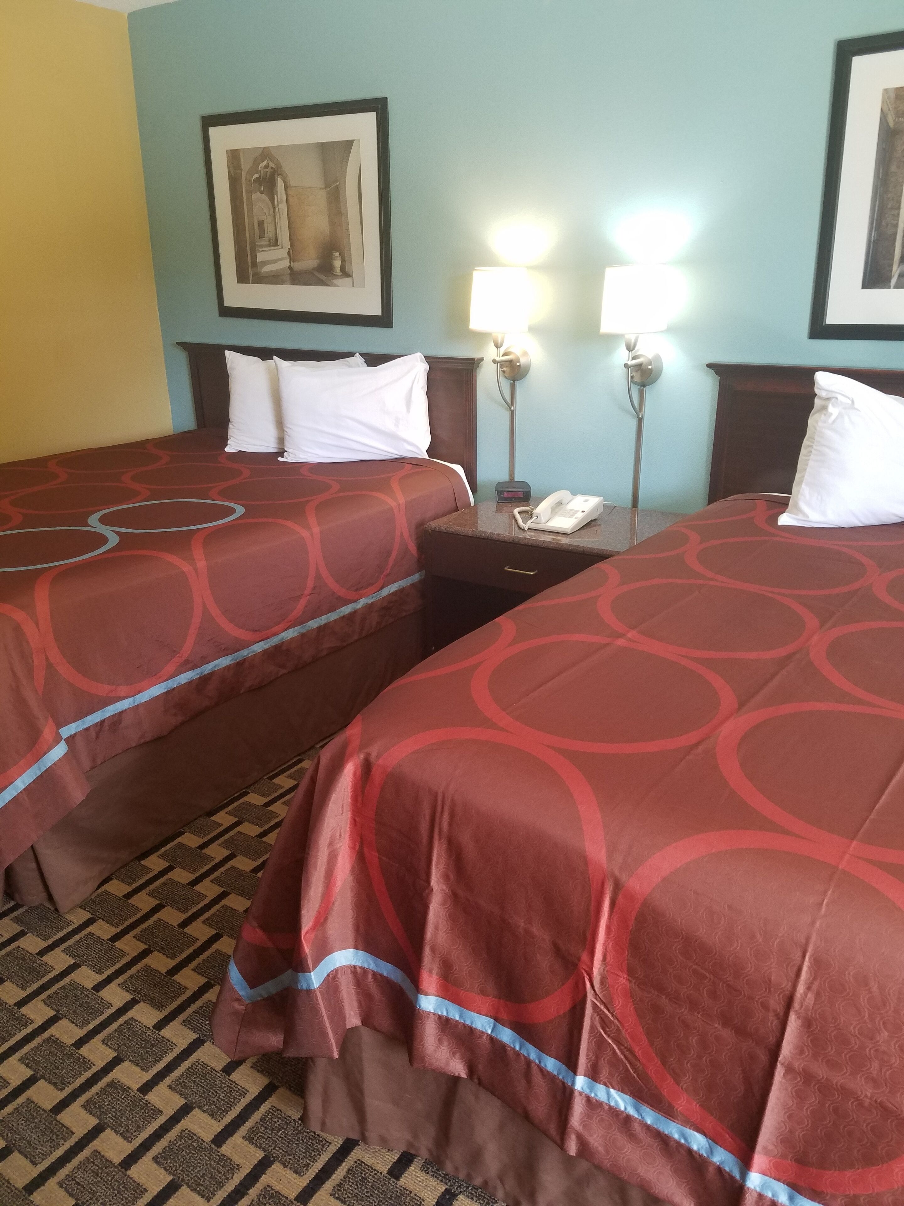 Ett hotellrum med två sängar, var och en med ett rött överkast och vita kuddar. Det finns ett nattduksbord med en lampa och en telefon på. Väggarna är målade i ljusblått och gult, och det hänger en inramad tavla på väggen.