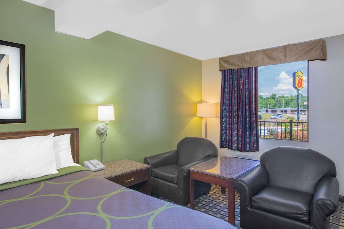 A hotel room with a bed, two chairs, a small table, a lamp, and a window with a view of a parking lot and a sign.