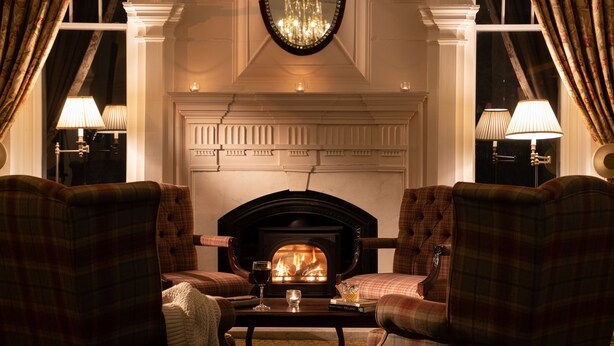 A cozy room with a fireplace, two plaid armchairs, a table with a glass of wine, and lamps on either side.