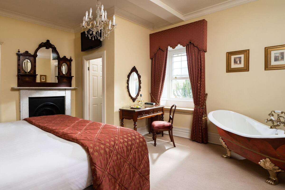 A bedroom with a bed, a desk, a chair, a fireplace, a mirror, a chandelier, a window with curtains, and a bathtub.