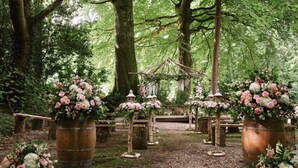A forest wedding setup with wooden barrels as tables, adorned with pink and white flowers.