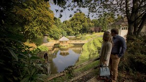 A couple standing by a pond with a wooden bridge, surrounded by lush greenery and a house in the background.