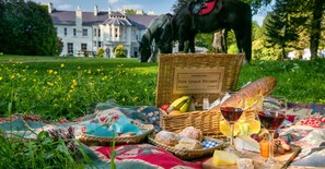 A picnic setup with a basket labeled "Veuve Clicquot Ponsardin," a bottle of wine, bread, cheese, and fruit on a blanket in a grassy area with a horse statue and a building in the background. - Beech Hill Country House Hotel (Londonderry)