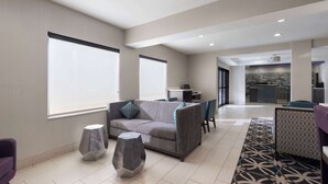 A modern hotel lobby with a grey sofa, two hexagonal stools, a patterned rug, and a view of the cityscape through large windows.