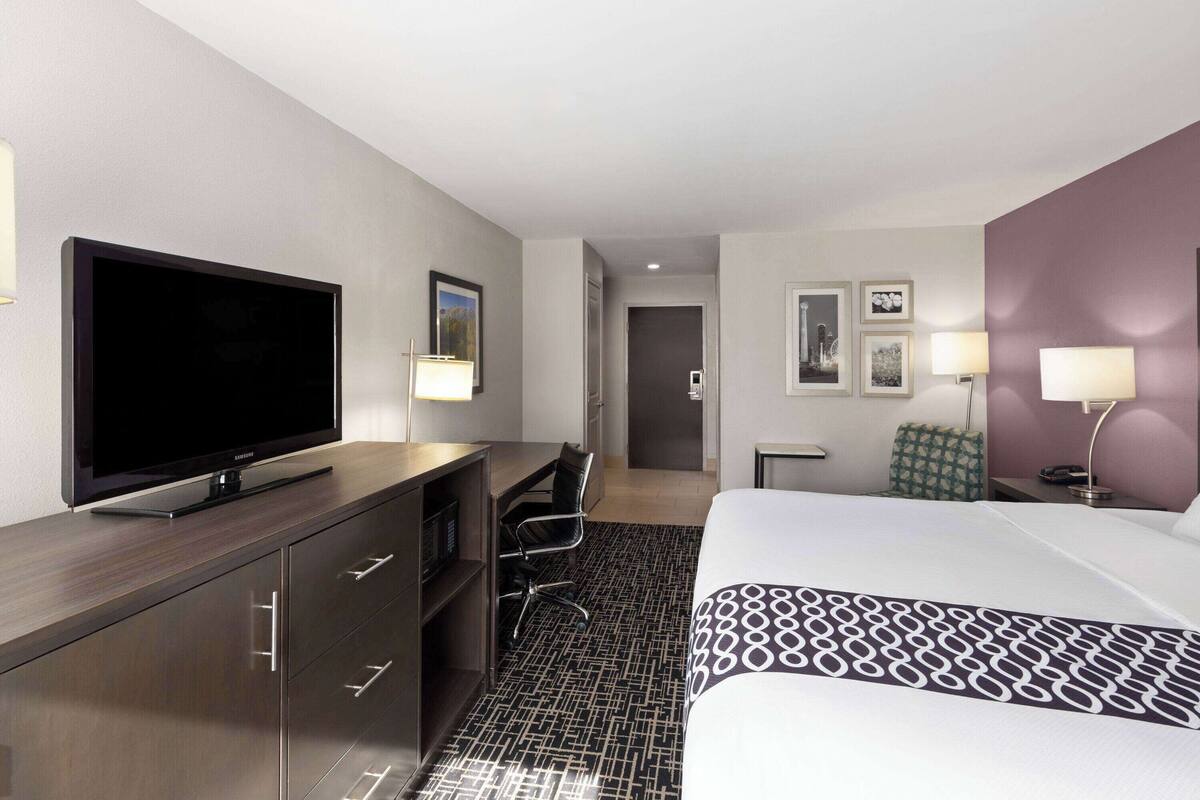 A hotel room with a bed, a desk with a TV, a chair, and a small table with lamps.