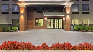 A modern hotel entrance with a covered walkway, glass doors, and a landscaped area with red flowers.