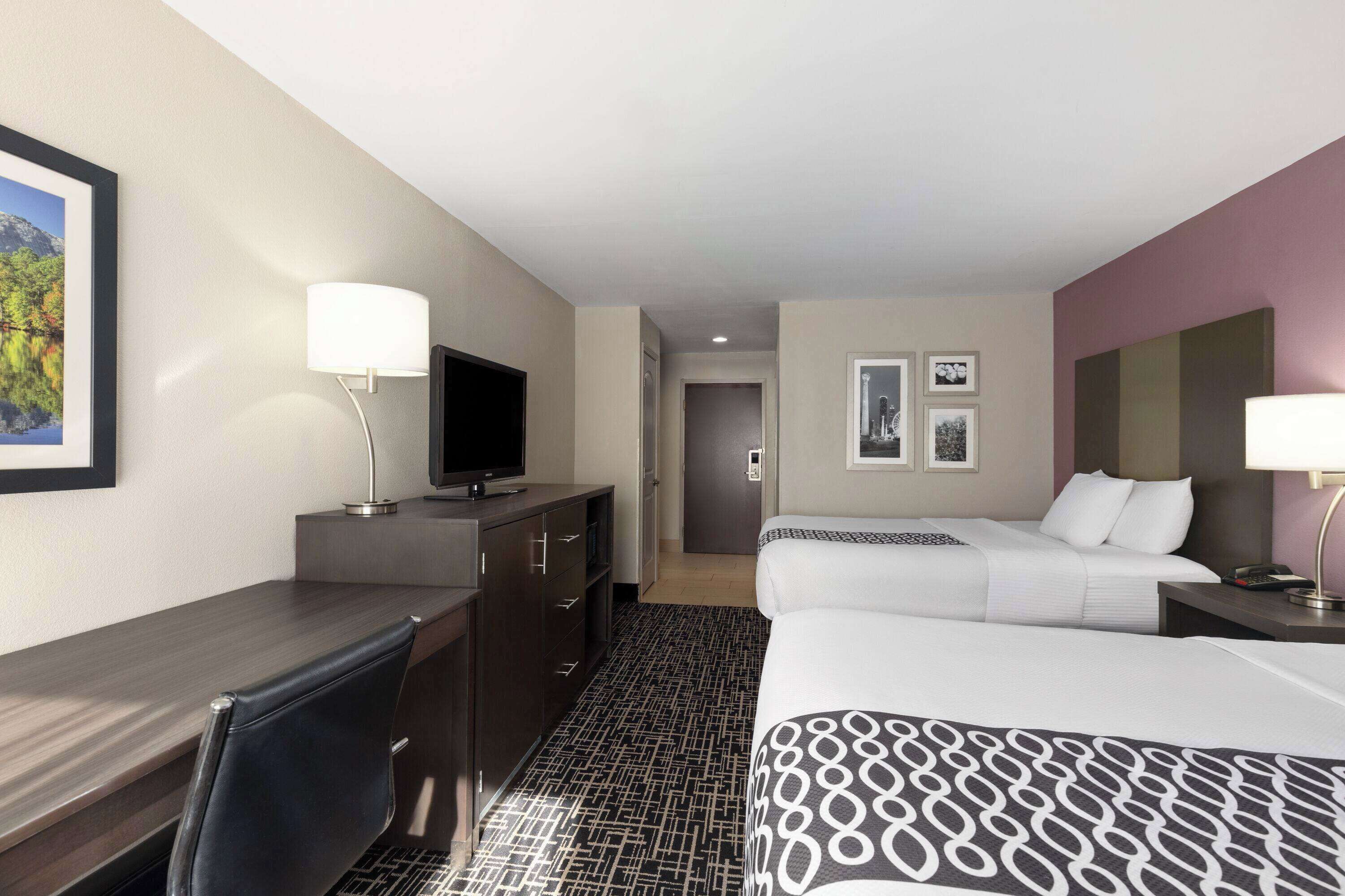 A hotel room with a desk, two beds, a television, and a bathroom visible in the background.