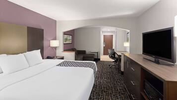 A hotel room with a large bed, a desk with a flat-screen TV, a chair, and a small table with a lamp.