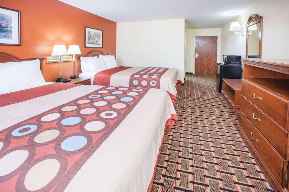 A hotel room with two beds, a wooden dresser, a patterned carpet, and a hallway leading to another room.