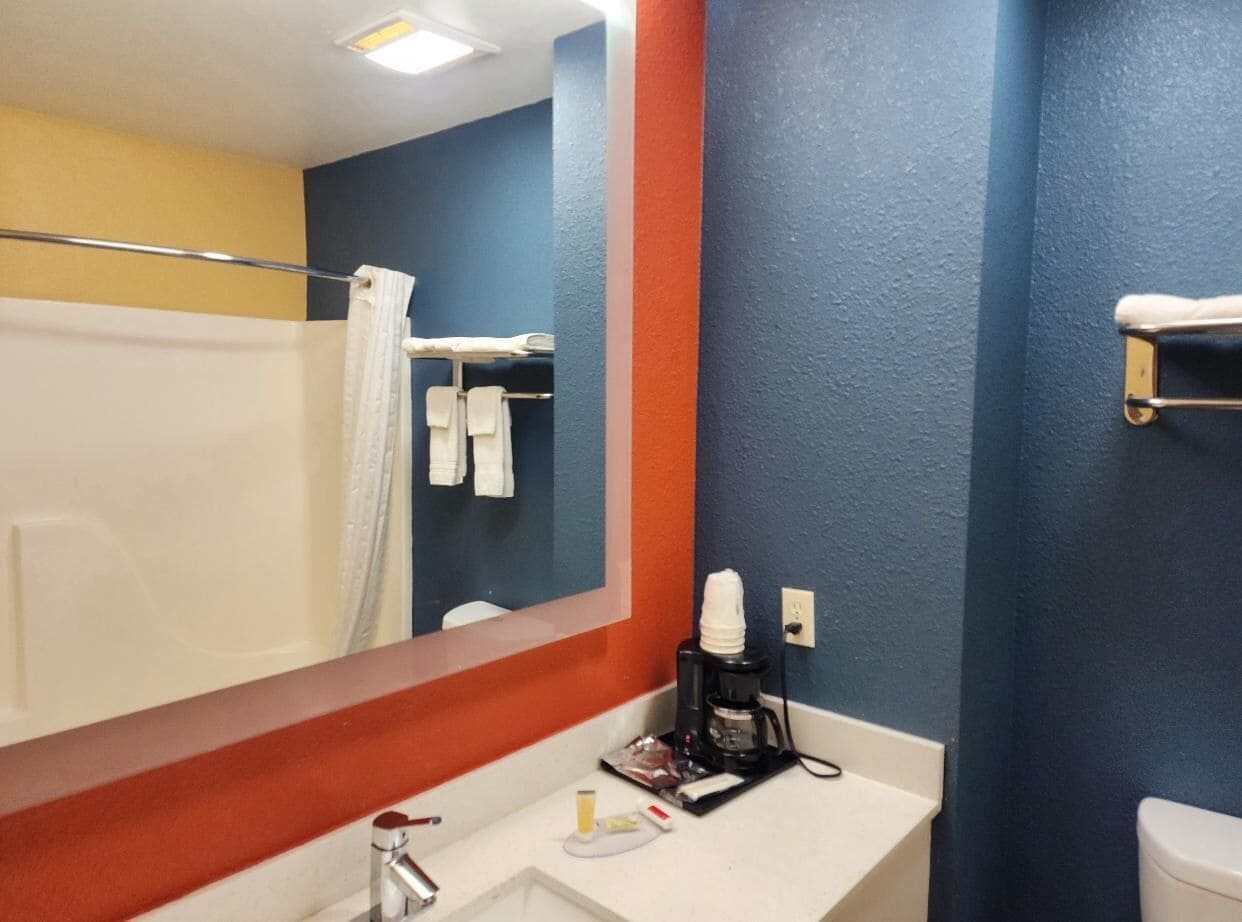 Un baño con lavamanos blanco, un espejo, una cafetera y toallas en un perchero.