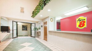 Zona de recepción de un hotel con una pared roja que muestra el logotipo 'Super 8', un mostrador de recepción, un reloj y una planta verde colgando del techo.