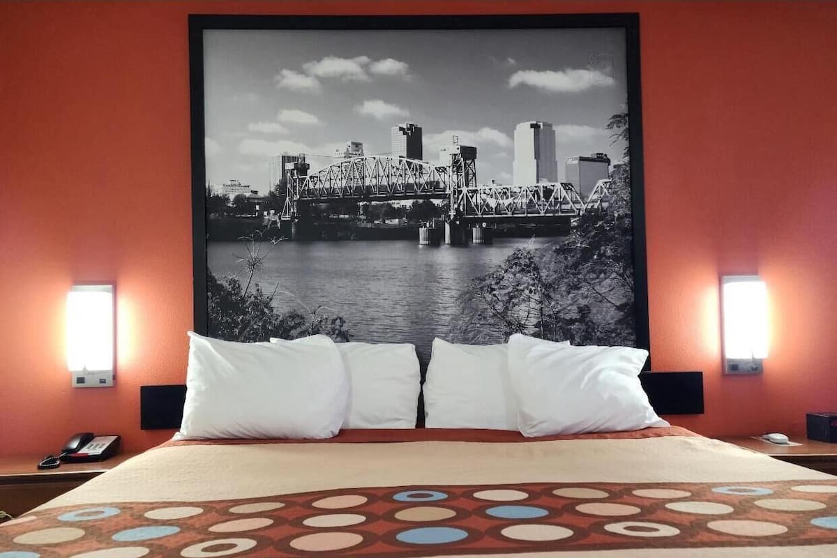 A hotel room with a large bed, two white pillows, a patterned bedspread, a cityscape view on the wall, and two bedside lamps.