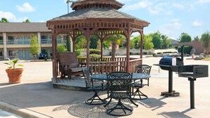 A gazebo with a wooden roof and metal chairs, a barbecue grill, and a mailbox.