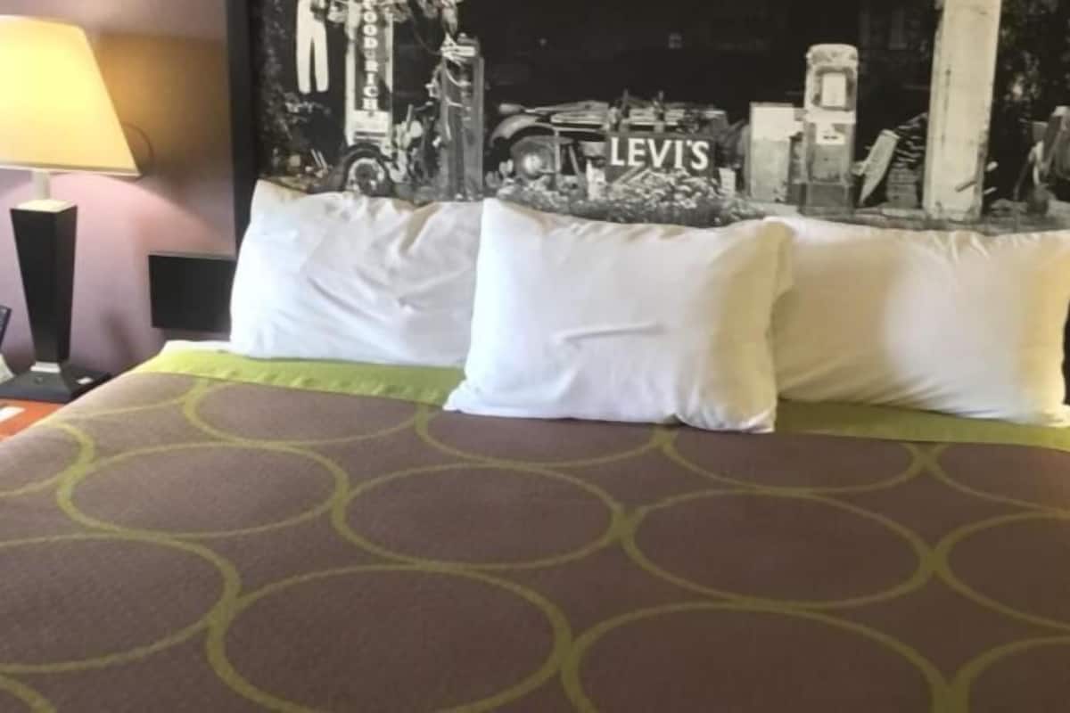 Hotelkamer met een bed met een beddegoed met een patroon, twee witte kussens, een nachtkastje met een lamp en een kunstwerk aan de muur waarop een stadsbeeld te zien is, met de tekst 'LEVIS'.