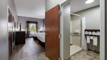 A hotel room with a bed, a desk, a chair, a window with curtains, a bathroom with a shower, and a towel rack.