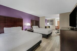 A hotel room with two beds, a desk, a chair, a television, and a wall with framed pictures.