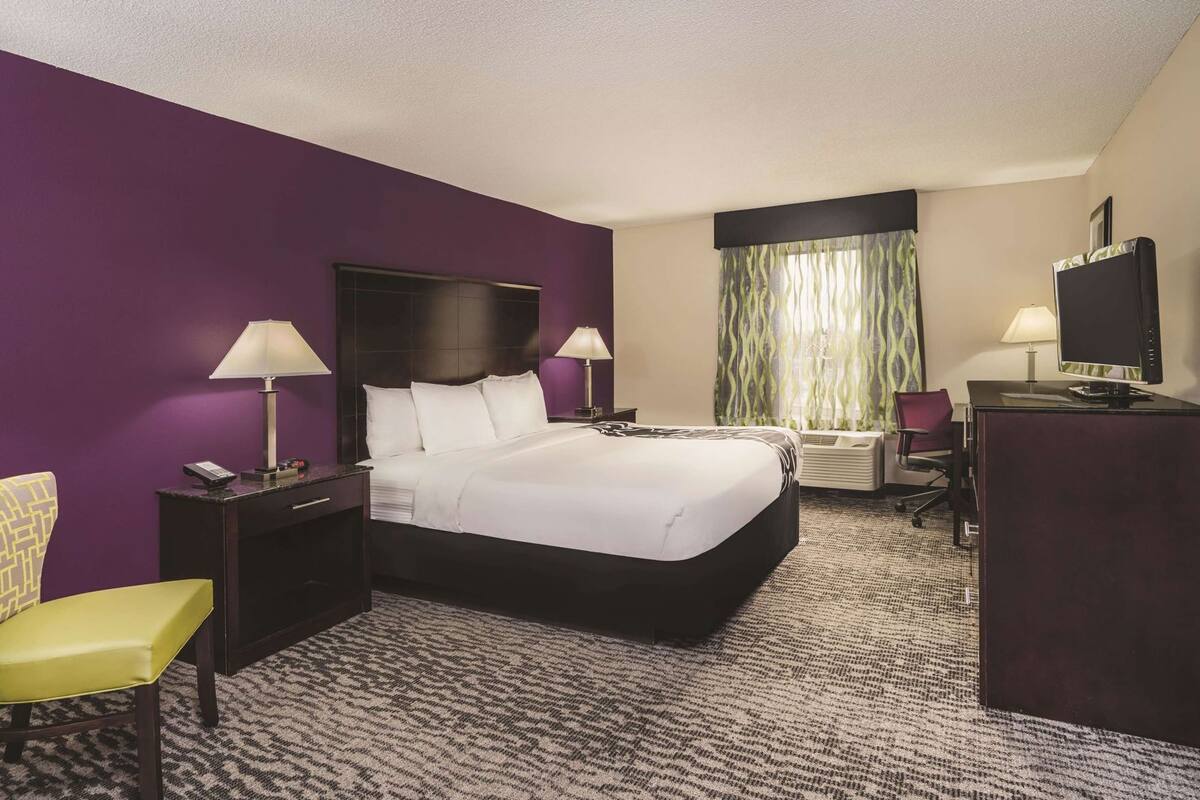 A hotel room with a bed, two bedside tables with lamps, a desk with a computer, a chair, and a large window with curtains.