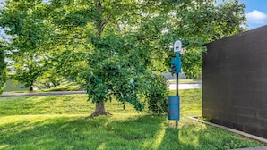 A pet waste station with a trash bin and a sign, located in a grassy area with trees and a brick wall in the background.