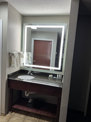 A bathroom with a dark granite countertop, a white sink, and a mirror with a lighted frame. There are towels hanging on a rack and a door visible in the mirror reflection. - La Quinta Inn & Suites by Wyndham Louisville East (Louisville)